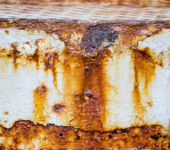 Detalle de superficie metálica blanca corroída con manchas de óxido marrón y naranja, mostrando deterioro y textura rugosa.